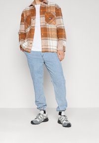 W kratkę pomarańczowa i biała koszula flanelowa na białym t-shircie, jasnoniebieskie dżinsy i szare buty sportowe z czarnymi akcentami.