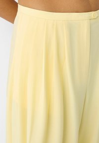 High-waisted pale yellow wide-leg pants with pleats and a button closure worn by a person with brown skin.