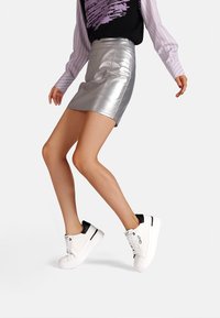 Sneakers bianchi con dettagli neri sul tallone, caratterizzati da una texture in pelle liscia, punta rotonda e suola piatta in gomma, indossati con una gonna metallizzata argento.