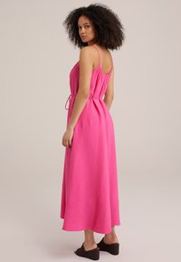Rosa, ärmlös maxi-klänning med texturerad tyg, åtsittande midja och justerbart bälte. Bärs med mörka klackslip-in, visad bakifrån.