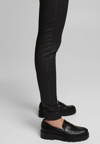 Personne portant un pantalon noir ajusté et des mocassins noirs à semelle épaisse, debout sur une surface lisse de couleur gris clair.