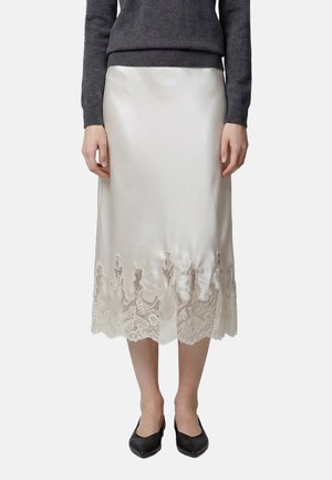 Frau trägt weißen Satin-Midirock mit Spitzenbesatz, dunkelgrauen Pullover und schwarze spitze Ballerinas, steht mit den Händen an den Seiten.