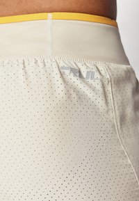 Vaaleanbeiget urheilushortsit, joissa on rei'itetty kangas, elastinen keltainen vyötärönauha ja pieni harmaa logo takana.