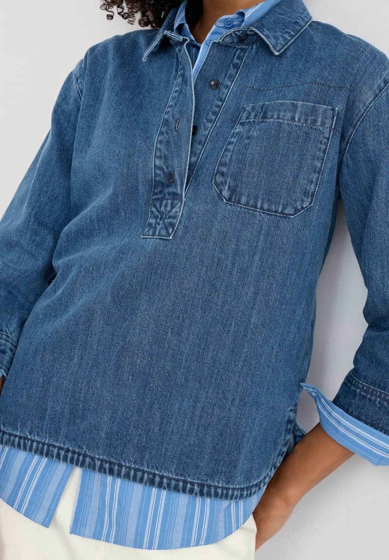 Pull-over en denim bleu foncé avec une patte de boutonnage, une poche avant et un ourlet rayé bleu contrastant. Tissu texturé avec manches longues.