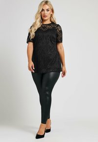 Top in pizzo nero con maniche corte, motivo floreale e scollatura rotonda, abbinato a leggings neri lucidi e tacchi neri.