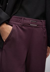 Ruka v kapse, oblečeny tmavě fialové kalhoty s elastickým pasem a černý top a bunda.