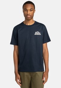 Navyblaue Baumwoll-T-Shirt mit kurzen Ärmeln und Rundhalsausschnitt. Mit einem weißen Grafiklogo auf der linken Brustseite. Angenehme, glatte Textur.