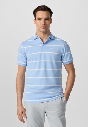 Jonge man met kort donker haar draagt een lichtblauw poloshirt met witte horizontale strepen en lichtgrijze broek, staat met één hand in de zak.