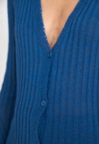 Blå ribbad kofta med V-ringning, knäppning framtill med en knapp och ett strukturerat stickat tyg.