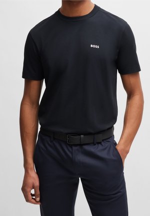 Mann trägt ein schwarzes "BOSS" Kurzarm-T-Shirt, das in navyblaue Hose mit schwarzem Gürtel gesteckt ist, steht mit einer Hand in der Tasche.