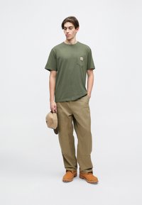 Olivově zelené triko s krátkým rukávem a kapsou na hrudi, spárováno s khaki kalhotami a hnědými dřeváky. Model drží béžovou čepici. Jednoduché pozadí.