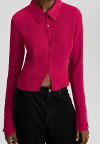 Camisa de manga larga fucsia con un acabado texturizado, botón en la parte delantera, cuello y un diseño corto que presenta una ligera abertura en el dobladillo.