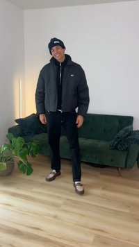 Ung mann som smiler, iført svart jakke, svarte bukser og lue, står i stuen med grønn sofa og potteplante på tregulv.