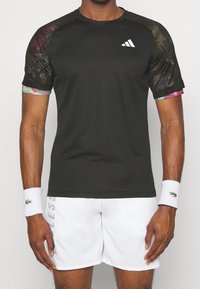 Camiseta atlética negra con mangas de patrón de malla que presentan acentos coloridos, combinada con pantalones cortos blancos y muñequeras blancas con logo.
