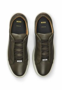 Gröna lädersneakers med en slät yta, med bruna snören, en vit gummisula och guldfärgade bokstäver "BOSS" på insidan och sidan.
