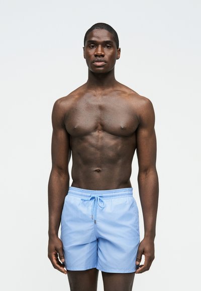 Νεαρός μυώδης άνδρας με ανοιχτό μπλε μαγιό, στέκεται μπροστά σε απλό φωτεινό φόντο, κοιτάζοντας μπροστά με ουδέτερη έκφραση.