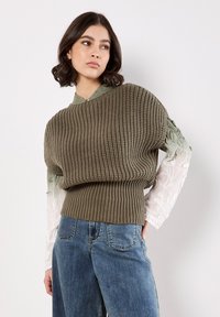 Sweter dzianinowy w oliwkowym zielonym kolorze z teksturowanym ciałem i prążkowanym dołem; posiada przezroczyste, białe haftowane rękawy. Połączony z jasnoniebieskimi dżinsami.