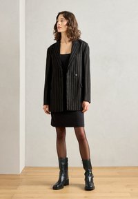 Czarna marynarka w paski o dopasowanym kroju, zapinana na jeden guzik, z dwiema przednimi kieszeniami, noszona na czarnej sukience i zestawiona z botkami do połowy łydki.