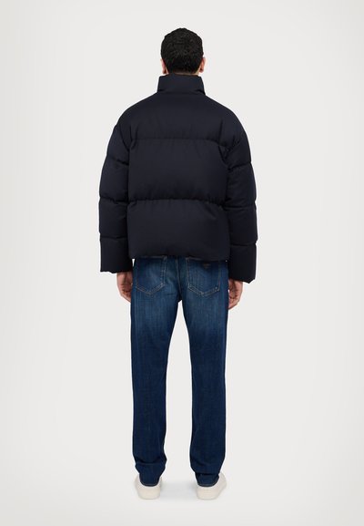 Navy puffer jacket with high collar, wide quilted design, and elastic cuffs, paired with dark blue straight-leg jeans and white sneakers.
