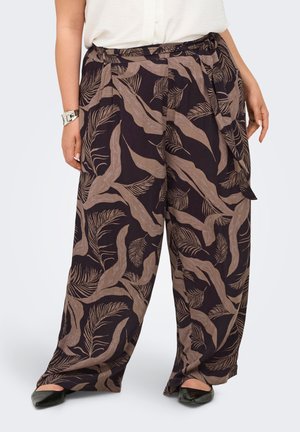 Femeie purtând pantaloni largi cu modele de frunze tropicale maro și o curea din material asortat, asortați cu pantofi negri și un top alb.