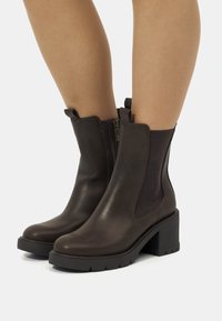 Bottes chevilles en cuir marron avec un talon bloque, fermeture éclair latérale et panneaux élastiques. Surface légèrement texturée et semelle en caoutchouc robuste.