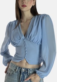 Hellblaue, transparente Langarmbluse mit tiefem V-Ausschnitt, gerafften Details und sechs Perlenknöpfen entlang der Vorderseite. Gekürzter Saum und glatte Textur.