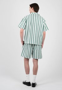 Jeune homme debout, portant une chemise à manches courtes rayée verticalement vert et blanc et un short assorti avec des chaussettes blanches et des chaussures noires, de dos.