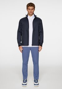 Marinblå vattentät jacka med hög krage, framdragkedja och två sidofickor, parat med en vit polo och ljusblå byxor.