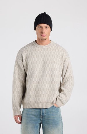 Mann trägt einen beigen strukturierten Strickpullover, blaue Jeans und eine schwarze Mütze, steht mit einer Hand in der Tasche vor einem schlichten Hintergrund.