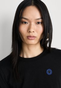 Black short-sleeve t-shirt featuring a blue embroidered smiley logo on the left chest, displayed against a neutral background.