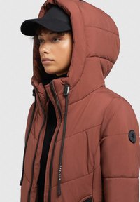 Brun oppustelig jakke med høj krave og hætte, struktureret stof, sorte snore og et rundt sort logo-patch på ærmet.