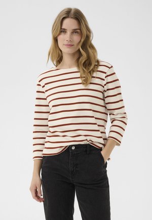 Femme aux cheveux blonds ondulés portant une chemise crème à rayures horizontales marron et un pantalon noir, debout avec une main dans la poche.