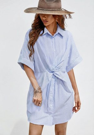 Frau trägt ein hellblau-weiß gestreiftes Hemdkleid mit kurzen Ärmeln und einem Knot in der Taille, accessoriert mit Armbändern und einem breitkrempigen Hut.