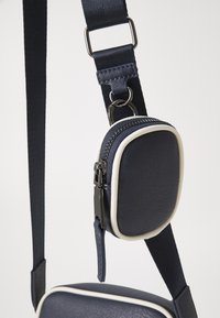 Borsa a tracolla blu navy con piping bianco, caratterizzata da una finitura testurizzata, tracolla regolabile e una piccola tasca con zip attaccata.
