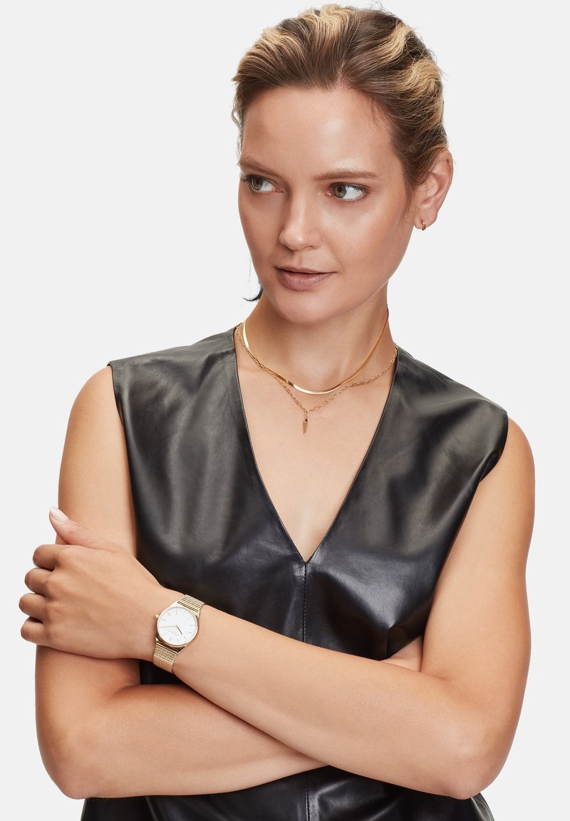 Gouden mesh horloge met een witte wijzerplaat, gedragen om een hand. De persoon draagt een zwart leren mouwloos topje en gelaagde gouden kettingen.