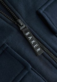 Jūras spēku audums ar teksturētu virsmu; satur melnu rāvējslēdzēju un melnu birku ar baltām burtiem, kas veido vārdu "BAKER."