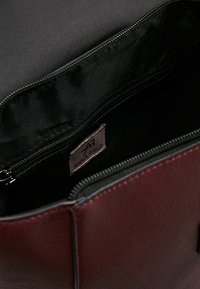 Borsa color bordeaux con esterno liscio, fodera in tessuto nero, tasca con cerniera e etichetta interna. Presenta una forma strutturata.