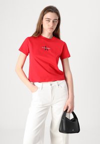 Punainen puuvillapaita, jossa on lyhyet hihat ja pyöreä pääntie, mustalla "Calvin Klein Jeans" -logolla, yhdistetty valkoisiin leveälahkeisiin housuihin ja mustaan laukkuun.