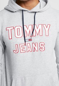 Sudadera gris con capucha y cordón, con texto "TOMMY JEANS" en rojo y blanco. Incluye un pequeño logo de bandera y un bolsillo canguro.