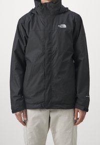 Uomo che indossa una giacca nera impermeabile The North Face con cappuccio e pantaloni cargo beige, in piedi contro uno sfondo semplice.