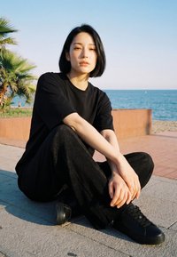 Young person in black clothing sits cross-legged on pavement near palm trees and ocean under clear sky.