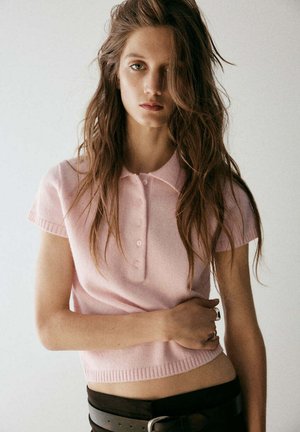 Joven mujer con largo cabello castaño lleva una camiseta polo rosa claro de manga corta y pantalones oscuros, de pie frente a un fondo claro y liso.