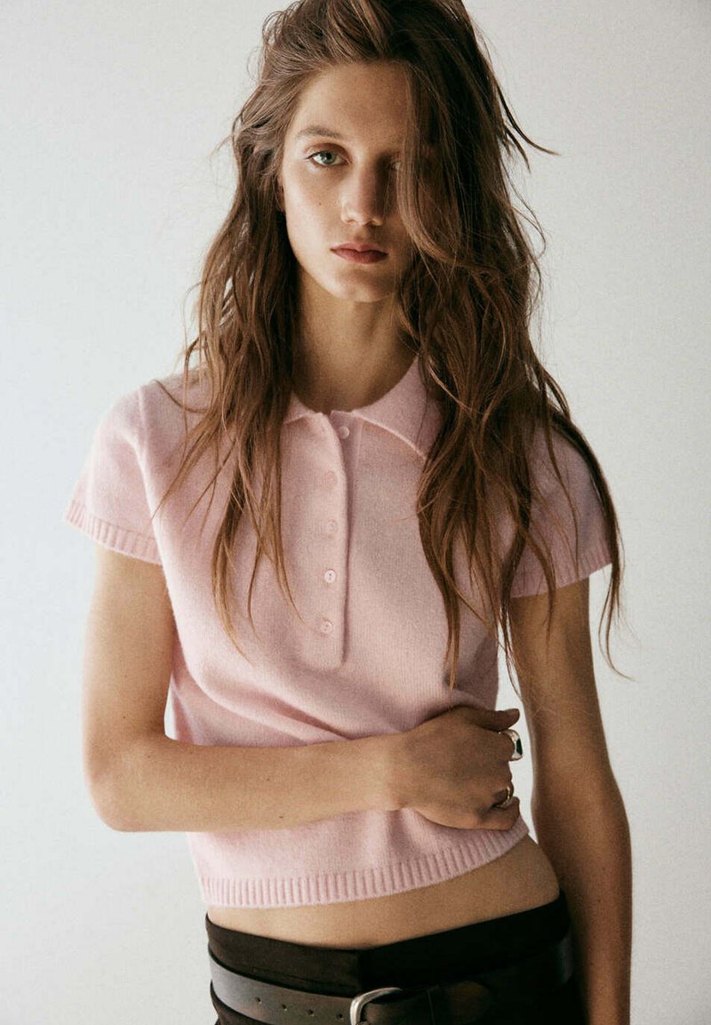Jeune femme aux longs cheveux bruns porte un polo rose clair à manches courtes et un pantalon foncé, debout devant un fond clair uni.