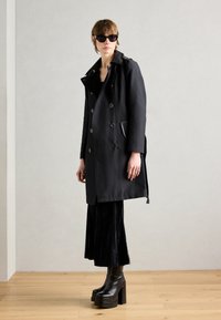 Manteau noir en trench avec un design à double boutonnage, grands revers, poches latérales, associé à une longue jupe noire et des bottes noires à talons épais.