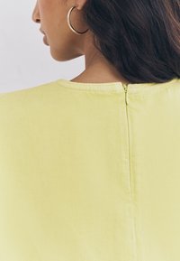 Indumento giallo con una texture liscia, presenta una chiusura posteriore con cerniera e una scollatura rotonda. Orecchino a cerchio argento visibile.