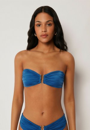 Frau mit langen dunklen Haaren, die einen blauen, gerafften, trägerlosen Bikini mit Metallverschluss-Details trägt und vor einem schlichten hellen Hintergrund steht.