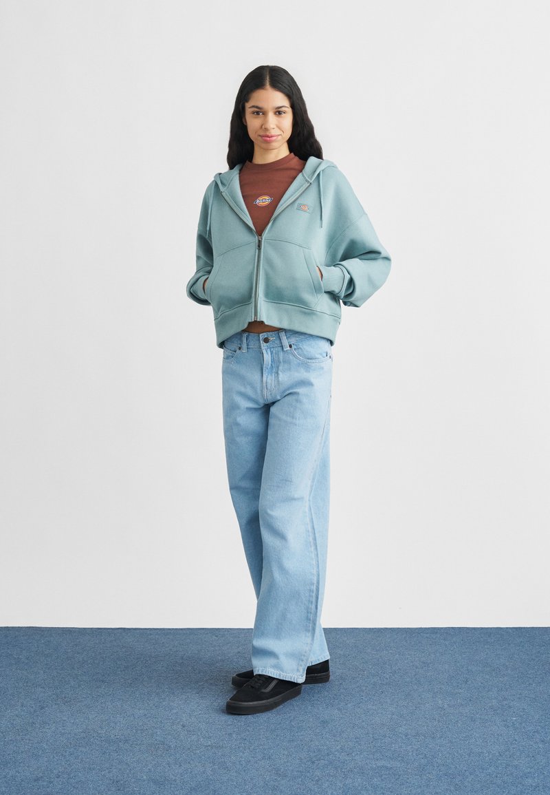 Jonge vrouw met lang donker haar, gekleed in een lichtblauwe hoodie met rits, een bruine t-shirt met logo, lichtblauwe jeans en zwarte sneakers, staand op een blauw tapijt.