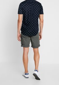 Uomo che indossa una camicia a maniche corte blu navy con quadrati bianchi, pantaloni corti grigi arrotolati e scarpe bianche New Balance, in piedi con la schiena rivolta verso la macchina fotografica.