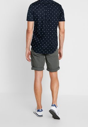 Man som bär en marinblå kortärmad skjorta med vita rutor, grå rullade shorts och vita New Balance-sneakers, står med ryggen mot kameran.