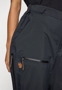 Technische zwarte broek met een gladde, matte textuur, met ritszakken met zwarte hardware en een bruine logo patch aan de zijkant.